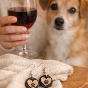 May include: A pair of round earrings with a black base and a wooden heart design. The wooden heart features a paw print. The earrings are displayed on a beige cloth, with a glass of red wine and a dog in the background.