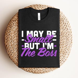 Könnte beinhalten: Schwarzes T-Shirt mit dem Text "I MAY BE Small - BUT I'M - The Boss" in weißer und lila Schrift. Das Shirt ist gefaltet und auf einem runden, gewebten, hellbraunen Platzset platziert.