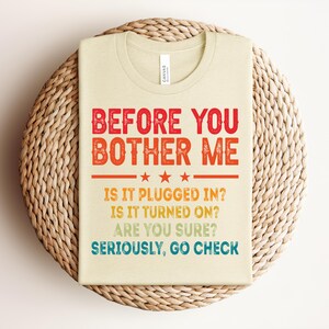 May include: A folded, light beige t-shirt with the text "BEFORE YOU BOTHER ME" in red and orange, followed by questions in yellow and blue. The shirt is resting on a woven, circular placemat.