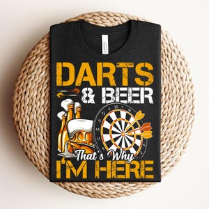 Può includere: T-shirt nero con grafica gialla. Il design include le parole "DARTS & BEER", un bersaglio per freccette, freccette e bottiglie di birra. È presente anche il testo "That's Why I'm Here".