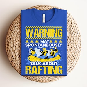 Può includere: T-shirt blu reale con grafica gialla e nera con la scritta "WARNING I MAY SPONTANEOUSLY TALK ABOUT RAFTING". La grafica raffigura un gommone giallo con tre persone che indossano giubbotti di salvataggio gialli. La maglietta è piegata su una superficie circolare intrecciata.
