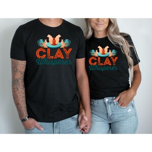 May include: Black t-shirts with the text "CLAY Whisperer" in orange and teal. The design includes pottery and hands. The shirts are worn by a couple, with the man having tattoos on his arm and wearing blue jeans.