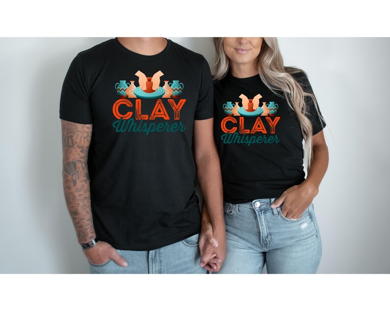 May include: Black t-shirts with the text "CLAY Whisperer" in orange and teal. The design includes pottery and hands. The shirts are worn by a couple, with the man having tattoos on his arm and wearing blue jeans.