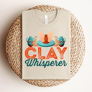 May include: Beige t-shirt with the text "CLAY Whisperer" in orange and teal. The design includes illustrations of hands shaping clay on a pottery wheel and two teal vases. The shirt is folded on a woven, circular placemat.