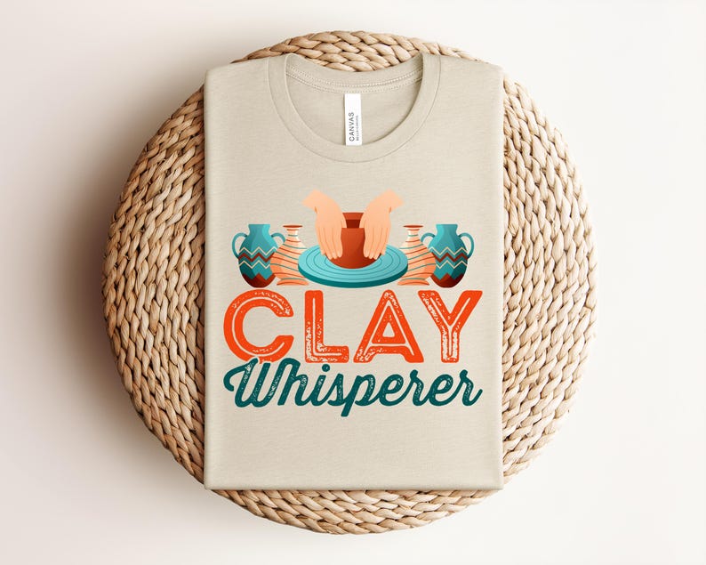 May include: Beige t-shirt with the text "CLAY Whisperer" in orange and teal. The design includes illustrations of hands shaping clay on a pottery wheel and two teal vases. The shirt is folded on a woven, circular placemat.