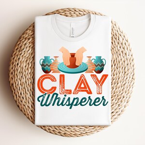 May include: White t-shirt with the text "CLAY Whisperer" in orange and teal. The design includes illustrations of hands shaping clay on a pottery wheel, and decorative vases. The shirt is folded on a woven, circular surface.