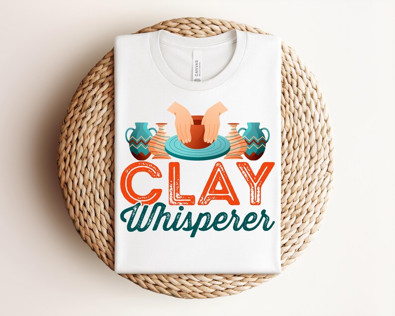 May include: White t-shirt with the text "CLAY Whisperer" in orange and teal. The design includes illustrations of hands shaping clay on a pottery wheel, and decorative vases. The shirt is folded on a woven, circular surface.