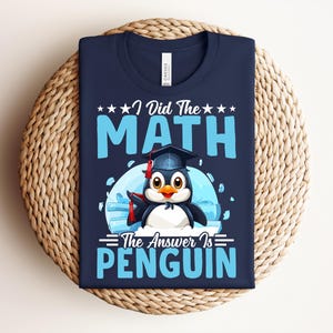 Op de afbeelding: Marineblauw T-shirt met de tekst "I Did The Math The Answer Is PENGUIN". De afbeelding toont een cartoonpinguïn met een afstudeerhoed en een diploma. De tekst en afbeelding zijn in tinten blauw en wit.