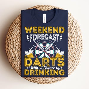Peut inclure: T-shirt bleu marine avec l'inscription "Weekend Forecast: Darts with a Chance of Drinking". Le motif comprend une cible de fléchettes, des fléchettes et des chopes de bière. Le t-shirt est plié sur un tapis tissé.