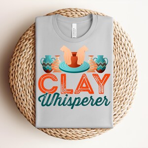 May include: A light gray t-shirt with the words "CLAY Whisperer" in orange and teal lettering. The shirt features a graphic of hands shaping clay on a pottery wheel, with two decorative vases on either side.