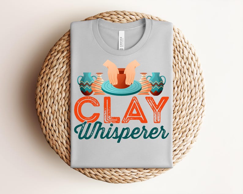 May include: A light gray t-shirt with the words "CLAY Whisperer" in orange and teal lettering. The shirt features a graphic of hands shaping clay on a pottery wheel, with two decorative vases on either side.