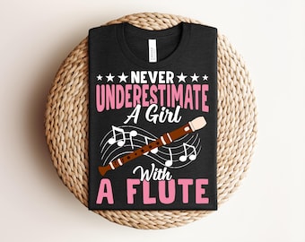 Peut inclure: T-shirt noir avec l'inscription "Never Underestimate A Girl With A Flute" en rose et blanc. Une flûte marron avec des accents blancs est représentée en dessous du texte, ainsi que des notes de musique.