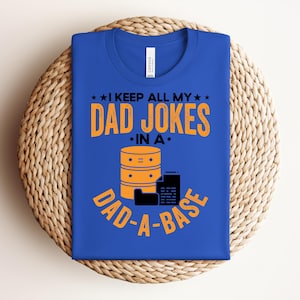 May include: Blue t-shirt with the text "I KEEP ALL MY DAD JOKES IN A DAD-A-BASE" in orange. The design includes a graphic of a server and a computer. The shirt is folded on a woven mat.