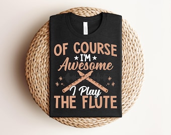 Peut inclure: T-shirt noir avec l'inscription "OF COURSE I'M Awesome I PLAY THE FLUTE" en beige. Le motif comprend des illustrations de flûtes et de notes de musique. Le t-shirt est présenté sur un dessous de plat circulaire tressé.