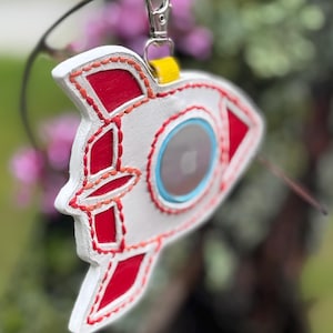 May include: White and red rocket-shaped keychain with a silver clasp and yellow strap. The rocket has a circular window with a light blue border. The design is accented with red stitching.