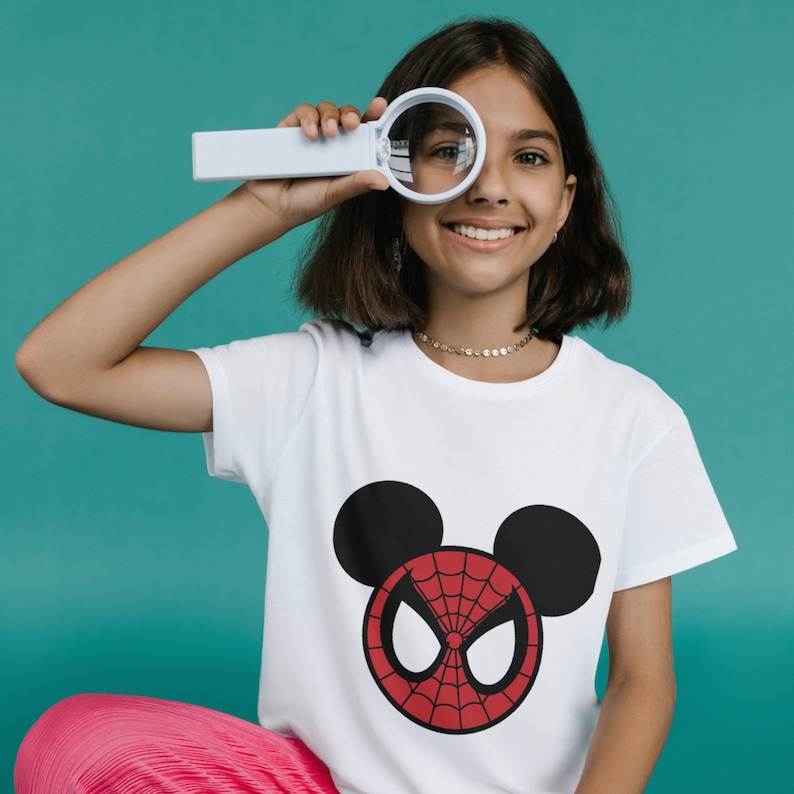 May include: A white t-shirt featuring a red and black Spider-Man graphic in the shape of Mickey Mouse's head. The shirt is being worn by a person with long brown hair.