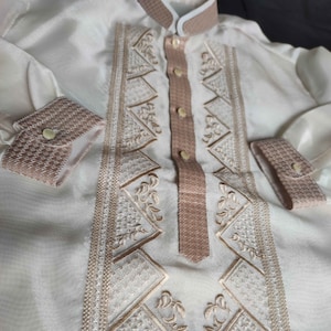 Puede incluir: Una camisa blanca de manga larga con un diseño bordado marrón. El bordado presenta formas geométricas y motivos florales.