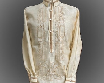 May include: A long-sleeved, cream-colored shirt with intricate embroidery in a lighter shade of brown. The shirt has a mandarin collar and a button-up front.