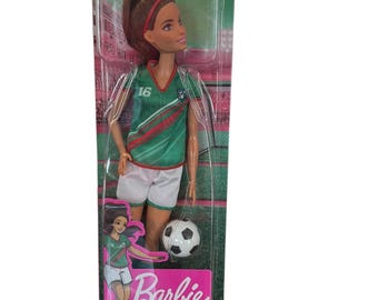 May include: A Barbie doll in a soccer uniform, packaged in a clear plastic box. The doll wears a green, red, and white uniform with the number 16. A soccer ball is included. The packaging reads "Barbie YOU CAN BE ANYTHING".
