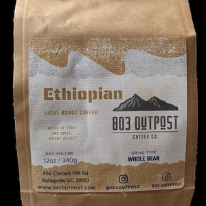 May include: A brown paper bag with a white label that says "Ethiopian Light Roast Coffee" and "803 Outpost Coffee Co.". The label also includes information about the coffee, such as the bag volume (12oz/340g), grind type (whole bean), and address of the company (456 Corbett Mill Rd., Bishopville SC 29010). The label also includes the company's website (www.803outpost.com) and social media handles (@803outpost and 803 Outpost).