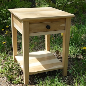May include: A light wood end table with a single drawer and a lower shelf. The drawer has a black knob. The table is sitting on a bed of green grass.