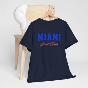 Op de afbeelding: Marineblauw t-shirt met het woord "MIAMI" in blauwe blokletters boven "Street Wear" in oranje letters. Een witte baseballpet ligt op een houten stoel.