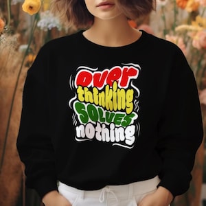 Puede incluir: Sudadera negra con el texto "Overthinking Solves Nothing" en rojo, amarillo, verde y blanco. La sudadera la lleva una persona. Fondo floral.
