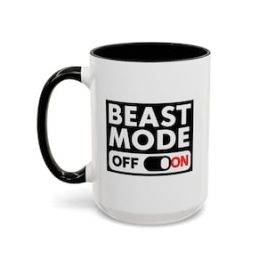 Puede incluir: Taza de cerámica blanca con interior, asa y borde negros. La taza presenta el texto "BEAST MODE" en letras negras en negrita, con "OFF" y "ON" en un gráfico de interruptor. El "ON" es rojo.