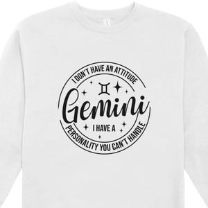 May include: White sweatshirt with a black Gemini zodiac design. The design includes the word "Gemini" in a script font, the Gemini symbol, and the phrases "I don't have an attitude, I have a personality you can't handle."
