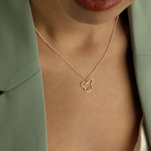 May include: A delicate gold-colored necklace featuring a minimalist mouse-shaped pendant. The pendant is a simple outline of the mouse's head and ears. The necklace is worn against a person's skin, with a green blazer in the background.