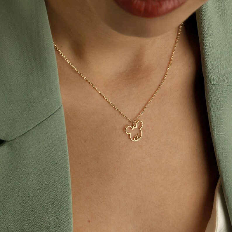 May include: A delicate gold-colored necklace featuring a minimalist mouse-shaped pendant. The pendant is a simple outline of the mouse's head and ears. The necklace is worn against a person's skin, with a green blazer in the background.