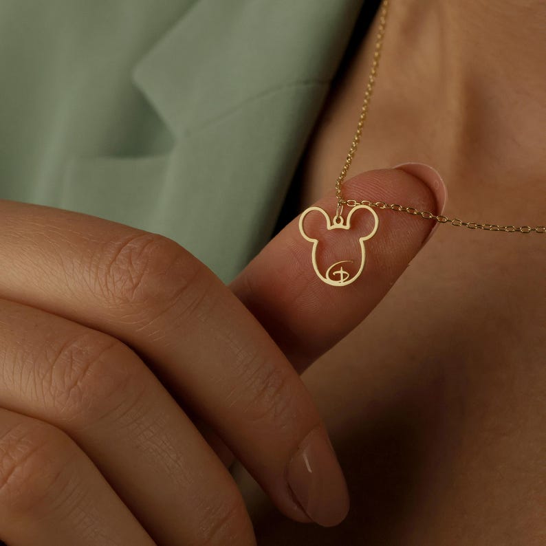 May include: A gold-colored pendant necklace featuring a Mickey Mouse silhouette. The pendant is a cutout design, showcasing the iconic character's head and face. The necklace is held by a finger, with a green background.