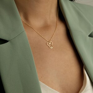 May include: A gold-colored necklace featuring a pendant in the shape of a mouse head. The pendant is a simple outline design. The necklace is worn against a person wearing a green blazer.