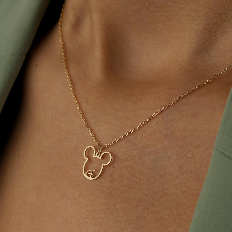 May include: A gold-colored necklace featuring a pendant in the shape of a cartoon mouse head. The pendant is a simple outline with a small detail for the face. The necklace chain is delicate and thin, and the background is a person wearing a green top.