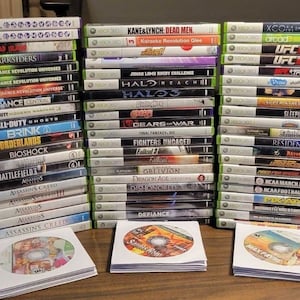May include: A collection of over 50 Xbox 360 video game cases stacked in three columns. The cases are arranged by game title and feature a variety of genres, including action, adventure, sports, and racing.