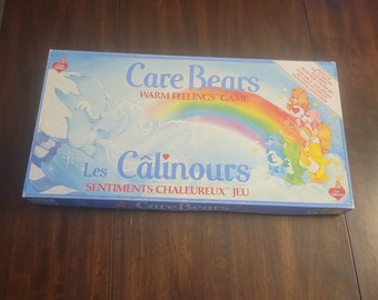 May include: Care Bears board game box with a blue and white background featuring a rainbow and Care Bears characters. The box is titled "Care Bears Warm Feelings Game" and "Les Calinours Sentiments Chaleureux Jeu".