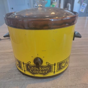 May include: A yellow Crockery Cooker slow cooker with a brown lid and a black knob. The cooker has a black and white design around the bottom with the words "Crockery Cooker" in a black banner.