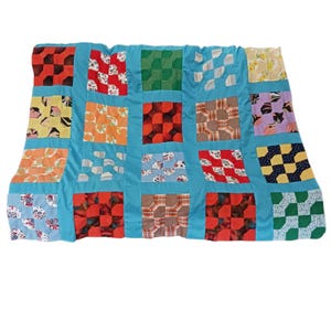 May include: A patchwork quilt featuring a grid of square fabric blocks in various colors and patterns, including red, orange, green, yellow, and floral prints. The blocks are separated by solid turquoise sashing, creating a structured design.