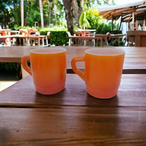 Könnte beinhalten: Zwei orangefarbene Tassen mit Henkeln stehen auf einem Holztisch. Die Tassen haben einen Farbverlauf, der von einem helleren Rosa am Boden zu einem leuchtenden Orange an der Oberseite übergeht. Der Hintergrund zeigt Sitzgelegenheiten im Freien.