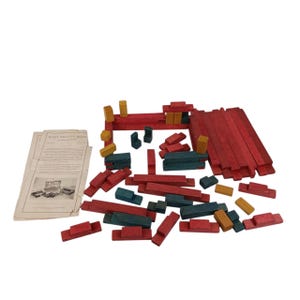 May include: A collection of wooden building blocks in red, blue-green, and yellow. The blocks are various shapes and sizes, including rectangular prisms and small cubes. A booklet with text and illustrations is also included.