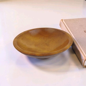 May include: A small, round, shallow wooden bowl with a smooth, polished finish and visible natural grain patterns in shades of golden brown. The bowl has a wide, flat rim and a curved interior basin, presented next to a beige hardcover book.