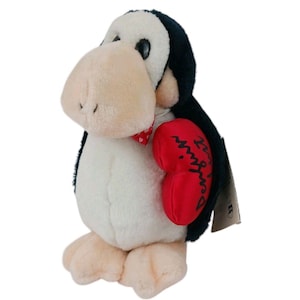 Pode incluir: Um brinquedo de pelúcia de pinguim com um grande bico e pés bege, corpo preto e branco e um acessório em forma de coração vermelho. O coração tem as palavras "Perfect Mingle" escritas. O pinguim usa uma gravata borboleta vermelha com bolinhas brancas.