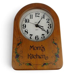 May include: An arched wall clock with a light brown wood grain finish. The white clock face features black Arabic numerals, black hands, and the text "Sunset" and "quartz". Below the clock face, the text "Mom's Kitchen" is printed in black, flanked by decorative green vine and purple grape illustrations.
