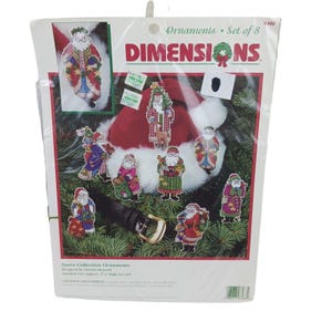 Puede incluir: Un kit de punto de cruz contado de la marca Dimensions para un juego de 8 adornos de la colección Santa. El empaque muestra ocho figuras distintas de Santa Claus con atuendos coloridos, un gorro rojo de Santa y un cinturón negro con hebilla.