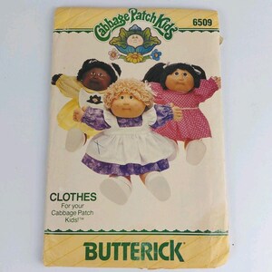 Puede incluir: Un sobre de patrón de costura Butterick, número 6509, para ropa de muñecas Cabbage Patch Kids. El frente muestra tres muñecas con varios atuendos, incluyendo un vestido amarillo, un vestido morado y blanco, y un vestido rosa con lunares.