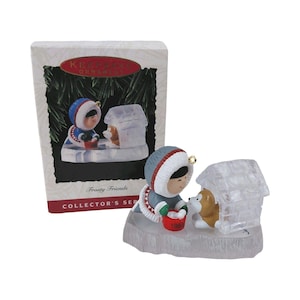 May include: A 1993 Hallmark Keepsake Ornament titled Frosty Friends featuring a figure in a grey and blue parka holding a red bucket next to a brown and white dog inside a translucent, block-patterned igloo structure on a textured base.