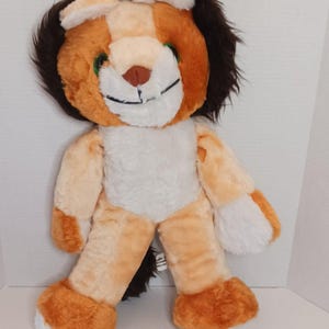 Op de afbeelding: Een pluche leeuw met een bruine manen, oranje en bruin vacht en een witte buik. De leeuw heeft een vriendelijke uitdrukking met groene ogen en een bruine neus. Het speelgoed is voor kinderen.
