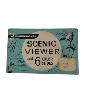 以下が含まれることがあります： ターコイズブルーの背景と円形デザインのヴィンテージ「Scenic Viewer」ボックス。「3 Dimensional」と「6 Color Slides」と書かれています。山とヤシの木のイラストが描かれています。ラベルには「Black Hills」と書かれています。
