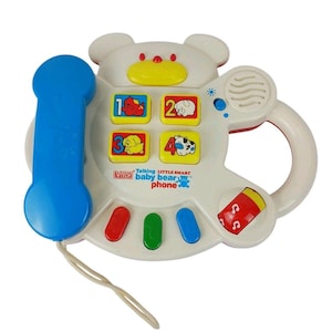 May include: A white VTech Talking Little Smart Baby Bear Phone toy with a blue handset attached by a cord. The device features a bear-shaped face, four yellow number buttons with animal illustrations, three colorful buttons, and a music note dial.