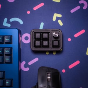 May include: A black keyboard with blue keys and a black control panel with four buttons and a knob. The keyboard and control panel are on a blue background with colorful geometric shapes.